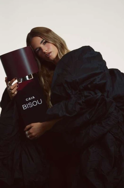 Brunette woman in ruffled black dress embraces a large perfume bottle, set against a pale background. The fragrance is 'CAIA BISOU'.