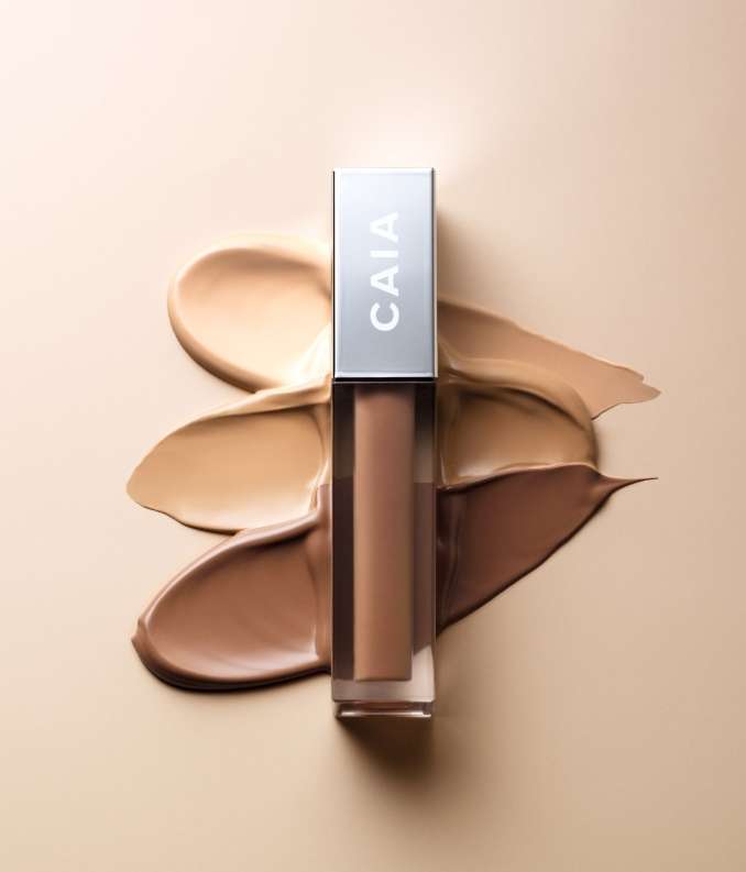 CAIA cosmetics concealer with silver lid and various shades of liquid foundation, on a smooth, light-beige surface in a studio setting.