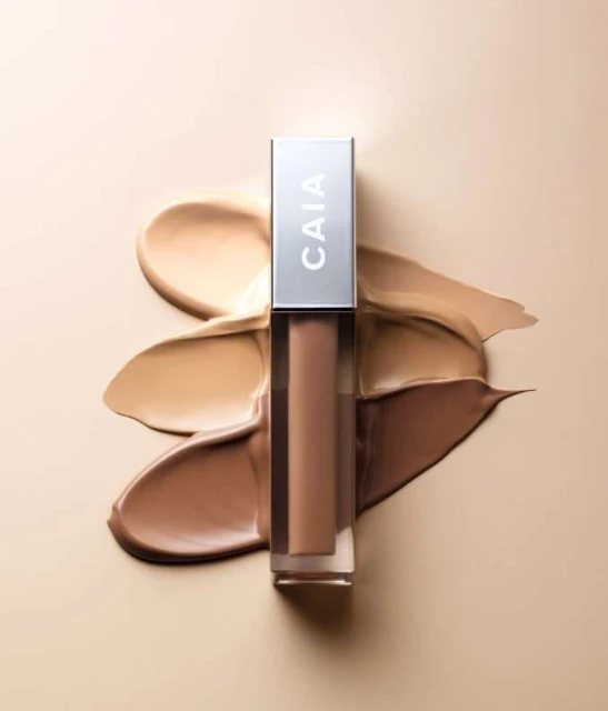 CAIA cosmetics concealer with silver lid and various shades of liquid foundation, on a smooth, light-beige surface in a studio setting.