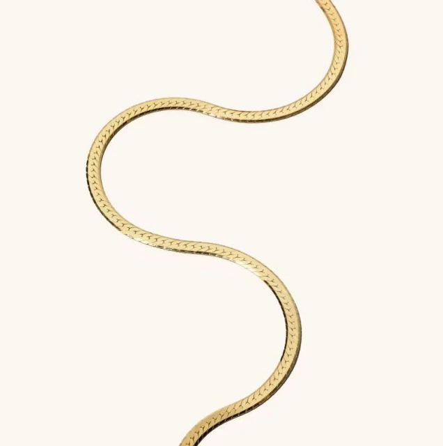Close-up studio shot of a single, smooth gold necklace in an s-shape on a seamless, light beige background creating a minimalist jewelry image.