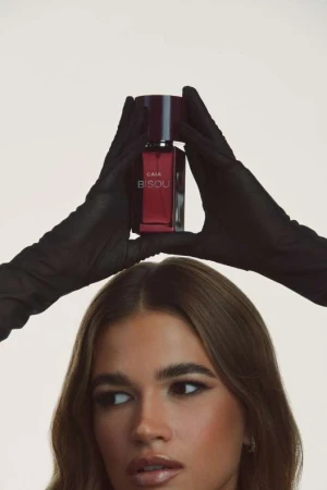 Close-up portrait of a woman holding a perfume bottle above her head wearing black gloves against a light background