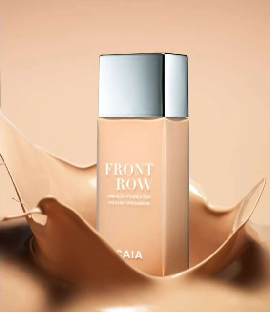 Beige foundation bottle with silver cap is surrounded by swirling liquid foundation against a light tan backdrop. Beauty shot.
