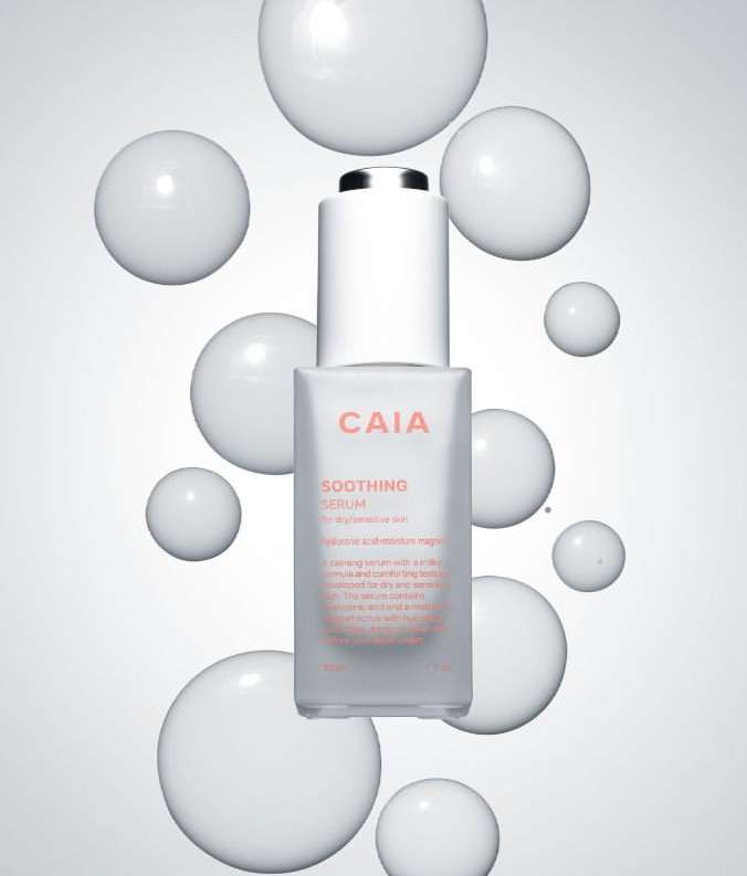 Bottle of CAIA Soothing Serum surrounded by glossy white spheres against a gradient white to grey background in a beauty product shot.