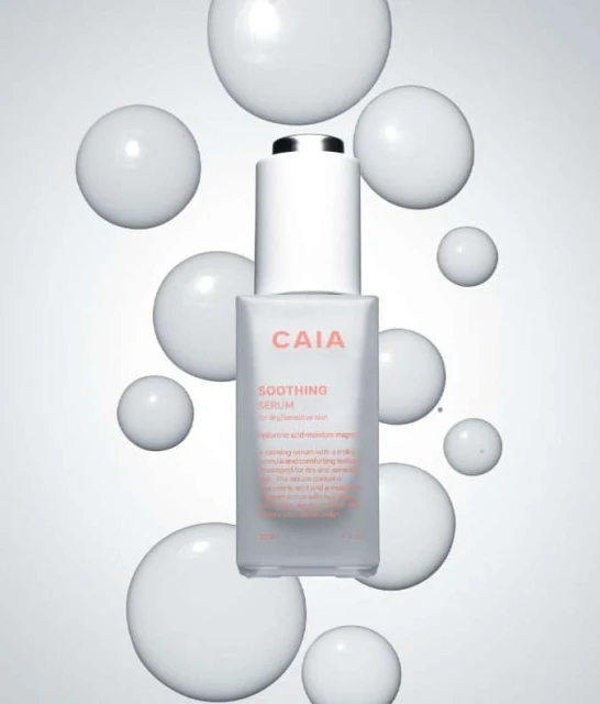 Bottle of CAIA Soothing Serum surrounded by glossy white spheres against a gradient white to grey background in a beauty product shot.