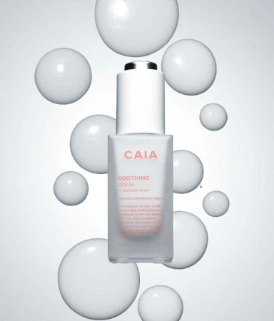 Bottle of CAIA Soothing Serum surrounded by glossy white spheres against a gradient white to grey background in a beauty product shot.