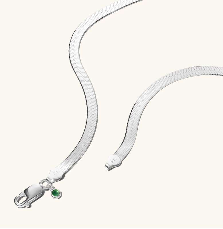 Elegant silver herringbone necklace with a lobster clasp and a green gemstone accent on a soft cream background. Minimalist jewelry shot.