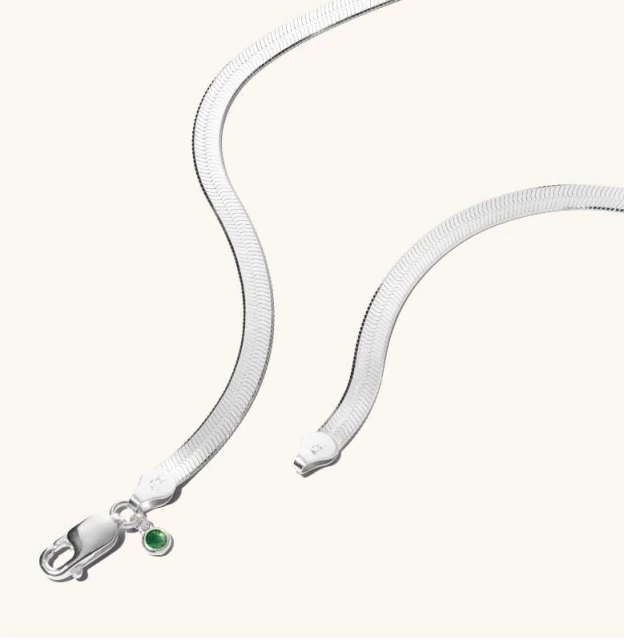 Elegant silver herringbone necklace with a lobster clasp and a green gemstone accent on a soft cream background. Minimalist jewelry shot.