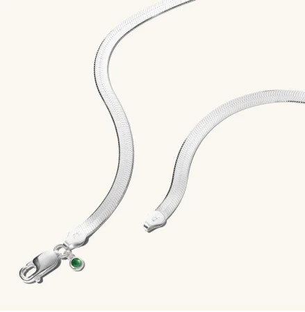 Elegant silver herringbone necklace with a lobster clasp and a green gemstone accent on a soft cream background. Minimalist jewelry shot.
