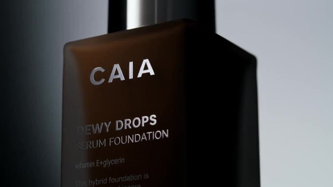 Close-up of CAIA Cosmetics 'Dewy Drops' serum foundation bottle. Product shot with soft lighting showcases brand name and product details.