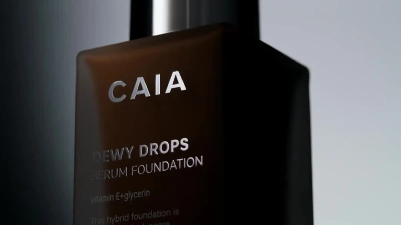Close-up of CAIA Cosmetics 'Dewy Drops' serum foundation bottle. Product shot with soft lighting showcases brand name and product details.