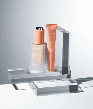Cosmetic product still life featuring CAIA brand lotion in a bottle and tube against a neutral backdrop and geometric acrylic display.