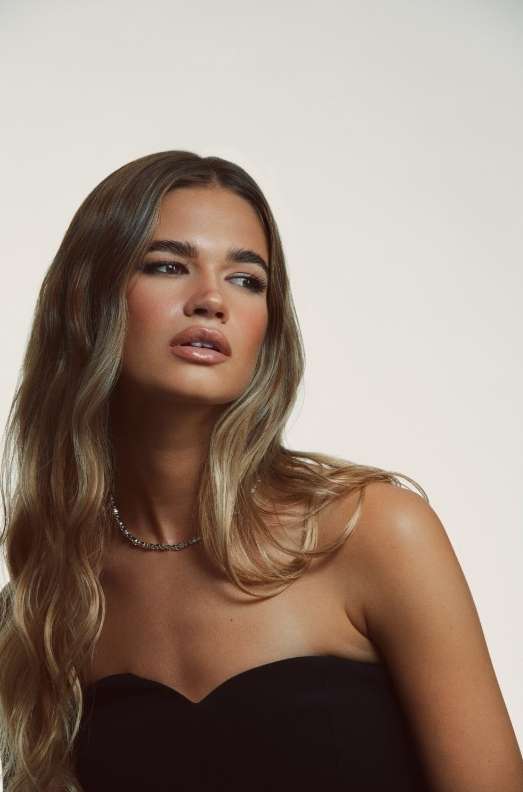 Portrait of a woman with long wavy brown hair wearing a black strapless top against a light background by Daniel Lindh