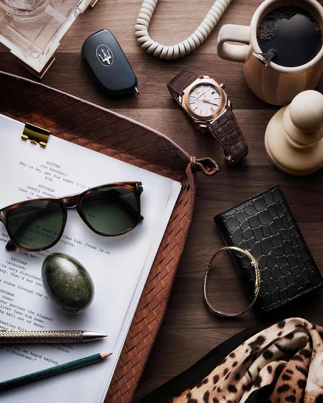 Overhead shot on a wooden surface featuring a script, sunglasses, watch, car key, coffee, wallet, bracelet, pens, and a scarf.