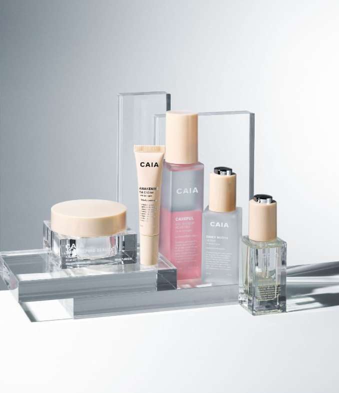 Collection of CAIA Cosmetics products including a tube, bottles, and jars, displayed on clear acrylic platforms with a light grey background.
