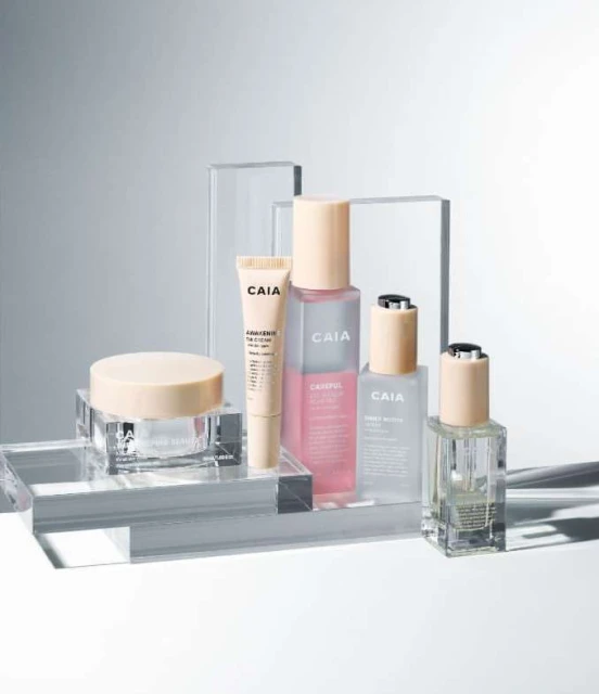 Collection of CAIA Cosmetics products including a tube, bottles, and jars, displayed on clear acrylic platforms with a light grey background.