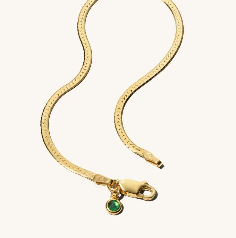 Close-up of a gold herringbone chain necklace with a green gem pendant, set against a plain, light background. Jewelry, fashion.