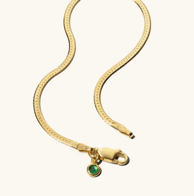 Close-up of a gold herringbone chain necklace with a green gem pendant, set against a plain, light background. Jewelry, fashion.