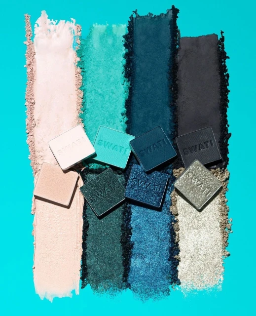 Eye shadow palette by SWATI Cosmetics with shades of beige, teal, and charcoal, arranged on a vivid turquoise background.