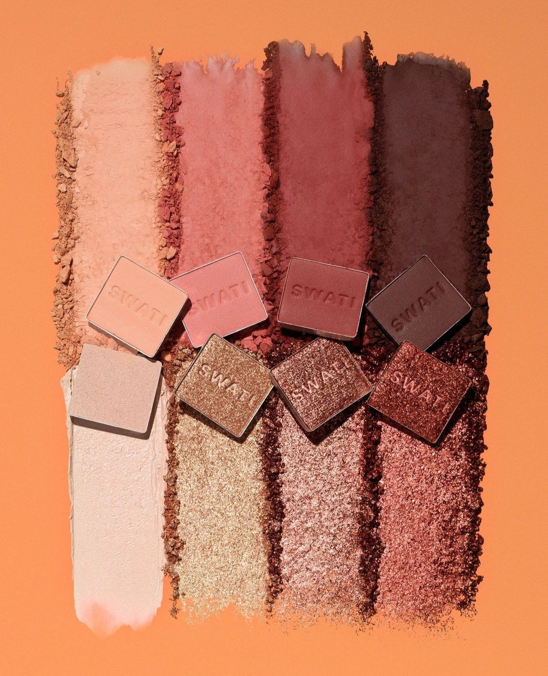 Eye shadow swatches of varying hues of brown and gold, with seven square sample containers arranged on top of the swatches.