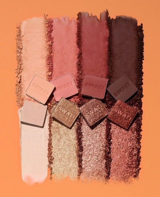 Eye shadow swatches of varying hues of brown and gold, with seven square sample containers arranged on top of the swatches.