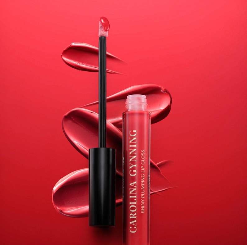 Close-up studio shot of Carolina Gynning brand red lip gloss, with applicator, set against a matching glossy red background.