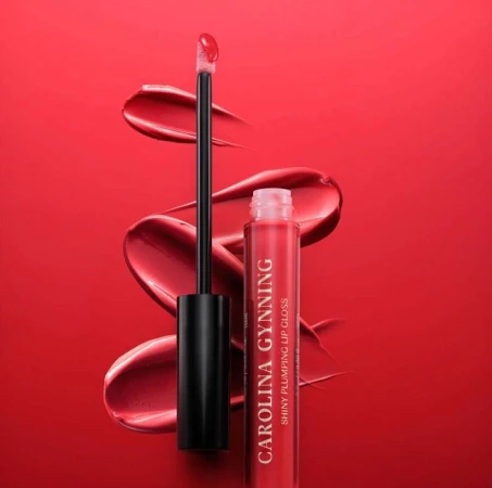 Close-up studio shot of Carolina Gynning brand red lip gloss, with applicator, set against a matching glossy red background.