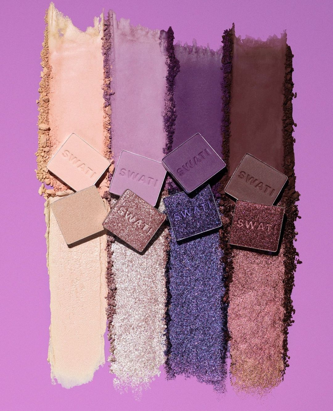Assortment of colorful eyeshadows and makeup arranged on a lilac background, showcasing different shades from light to dark.