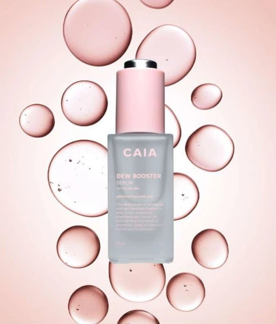 Elevated view of a 'CAIA Dew Booster' serum bottle with pink label and dropper against a light pink backdrop with translucent drops.