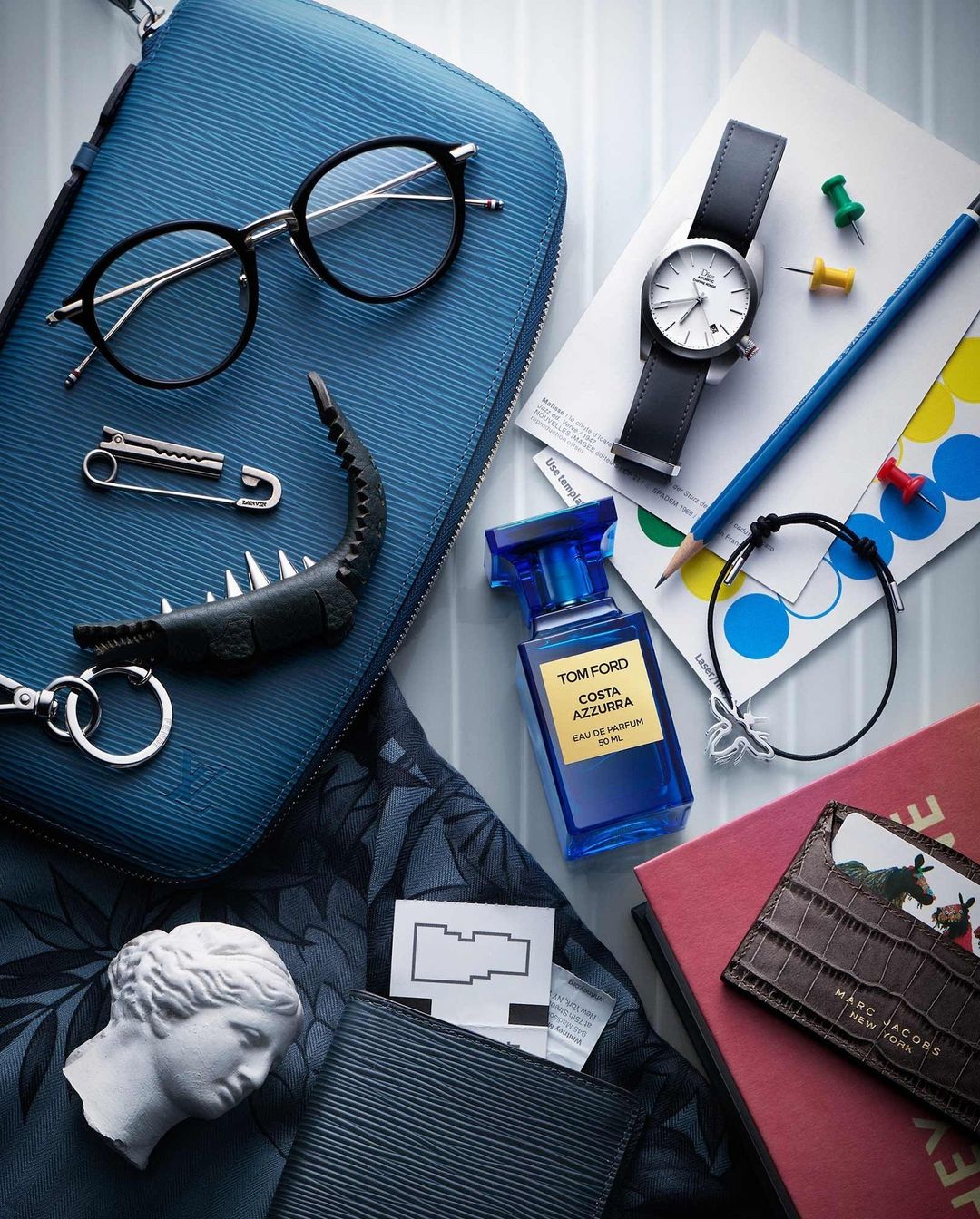 Overhead shot of fashion accessories including a blue leather pouch, glasses, watch, perfume, wallet, and miniature statue.