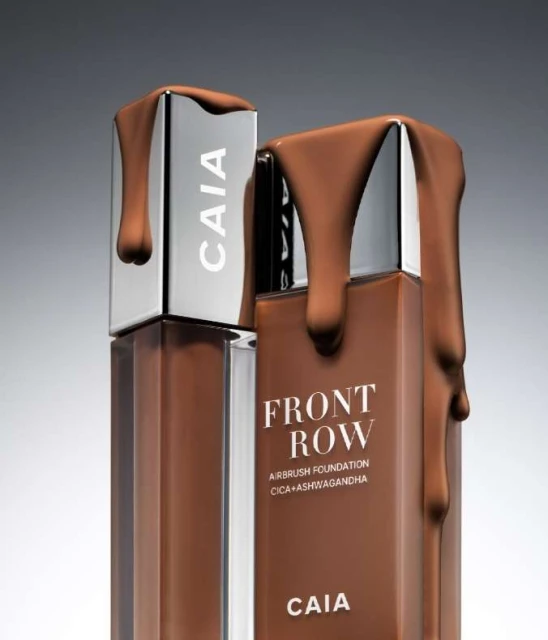 Two brown CAIA Front Row Airbrush Foundation bottles with silver tops and brown dripping foundation against a grey gradient background.
