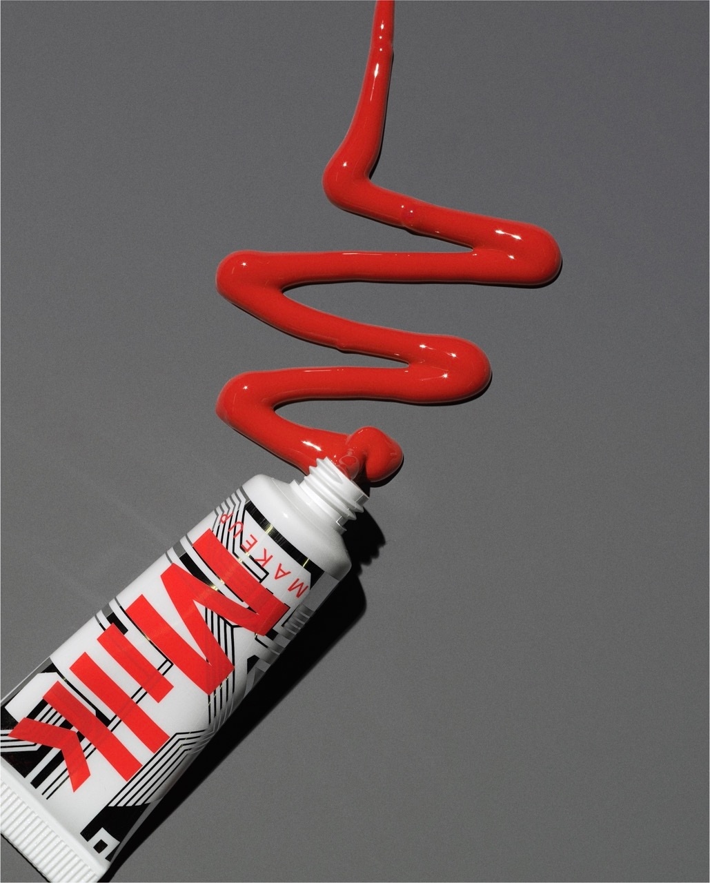Milk Makeup product shot with red liquid emerging from a white tube against a grey background, showcasing cosmetic application.