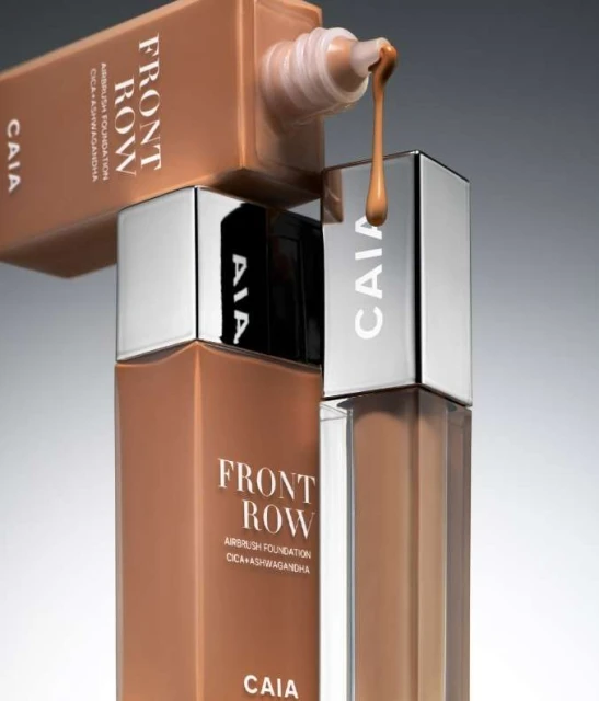 Close-up of CAIA Front Row Airbrush Foundation bottles, one tilted with liquid dripping, against a grey background.