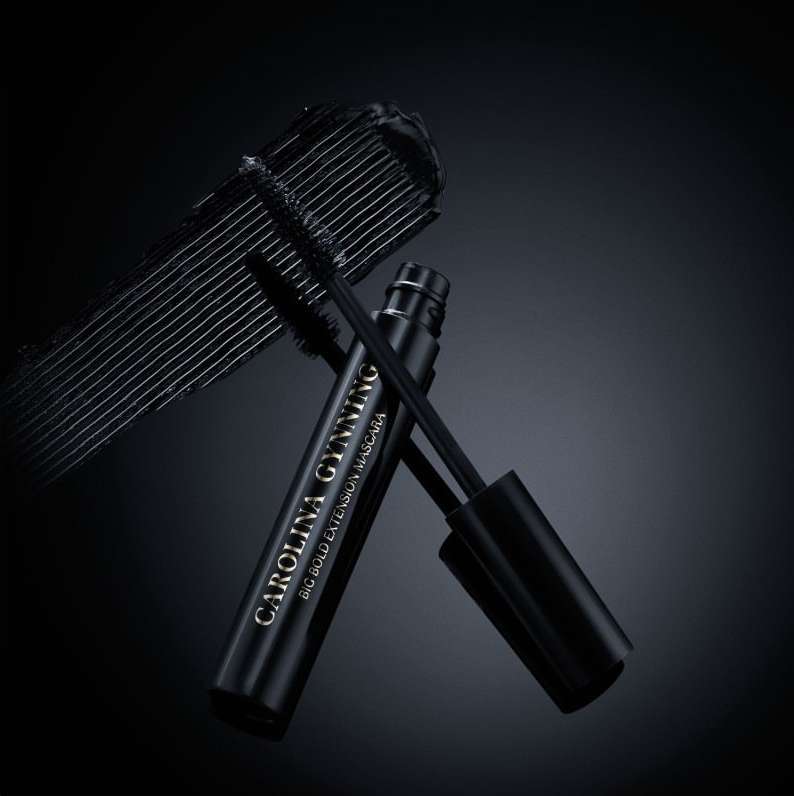 Close-up of Carolina Gynning Big Bold Extension Mascara tube and applicator wand with black mascara streak on a dark background.
