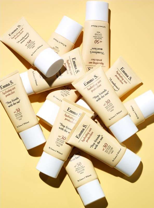 Several Emma S. hydrating sun protection tubes with SPF 30 and SPF 50 are displayed against a yellow background in a studio setting.