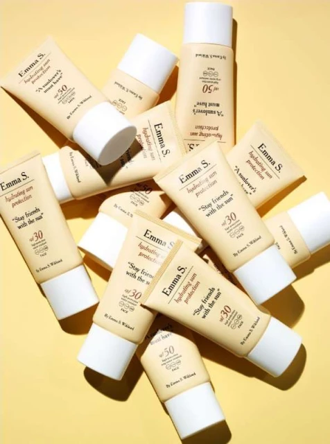 Several Emma S. hydrating sun protection tubes with SPF 30 and SPF 50 are displayed against a yellow background in a studio setting.