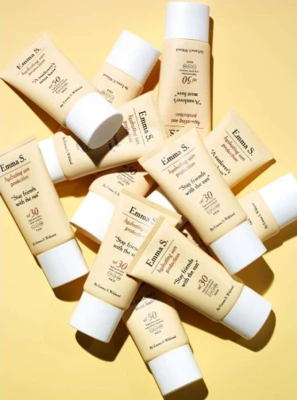Several Emma S. hydrating sun protection tubes with SPF 30 and SPF 50 are displayed against a yellow background in a studio setting.