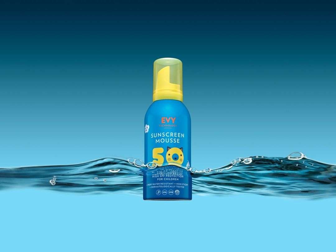 Blue bottle of sunscreen mousse for children is positioned in water with bubbles against a blue background in this product shot.