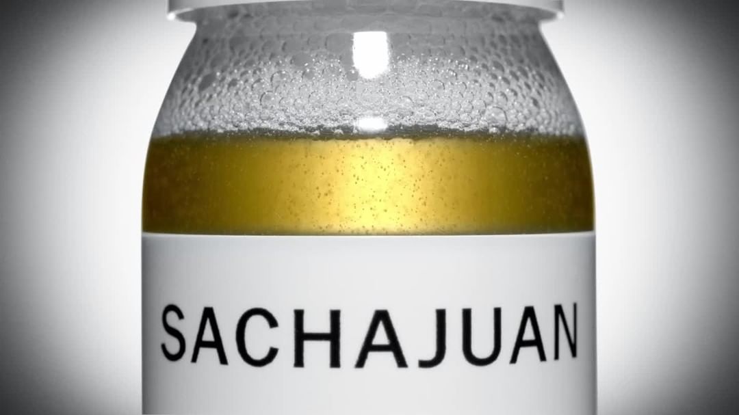 Close-up shot of a Sachajuan product in a clear glass jar with a white label, showcasing the liquid inside against a soft gray background.