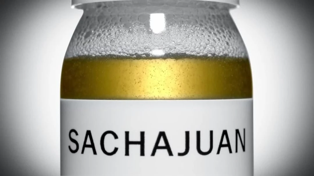Close-up shot of a Sachajuan product in a clear glass jar with a white label, showcasing the liquid inside against a soft gray background.
