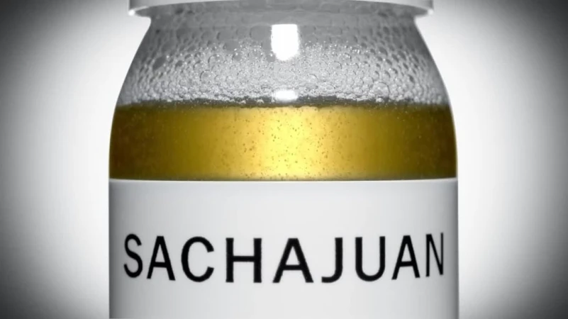 Close-up shot of a Sachajuan product in a clear glass jar with a white label, showcasing the liquid inside against a soft gray background.