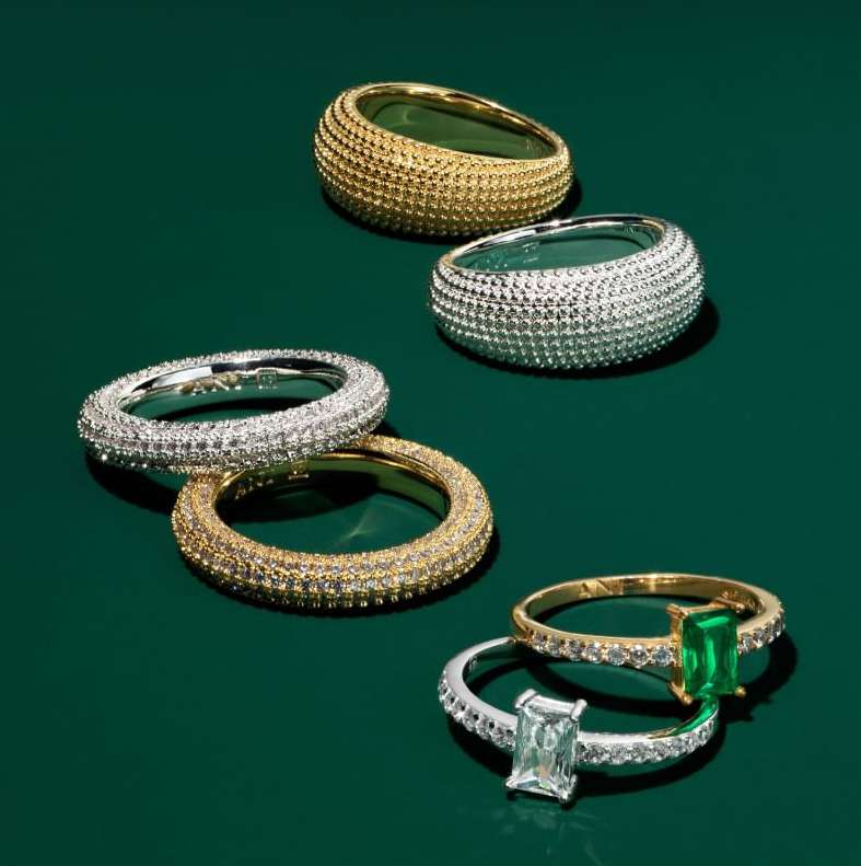 Close-up studio shot of gold and silver gemstone rings with diamond and emerald settings against a dark green background.