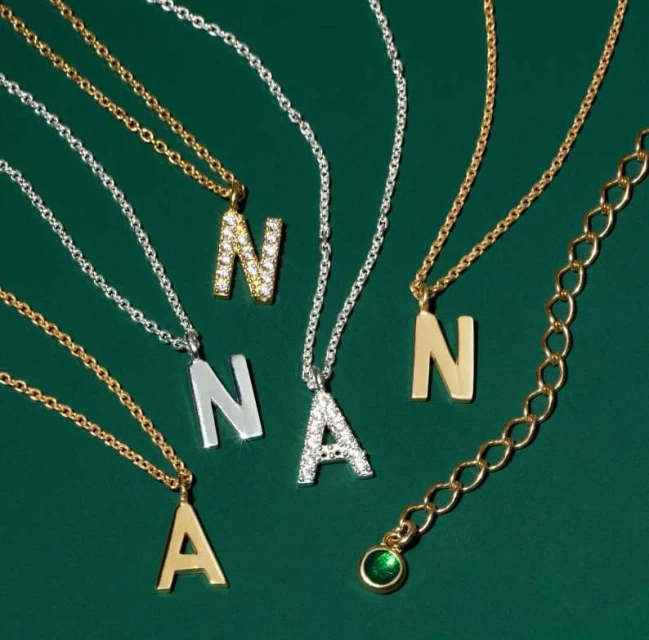 A close-up showcases gold and silver necklaces with initial pendants, set against a dark green background. Each letter is styled differently.