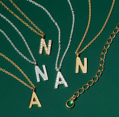A close-up showcases gold and silver necklaces with initial pendants, set against a dark green background. Each letter is styled differently.