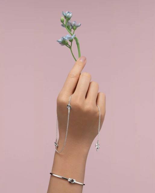 Close-up of a hand holding a sprig of light blue flowers, adorned with a silver necklace and matching bangle bracelet against pink backdrop.