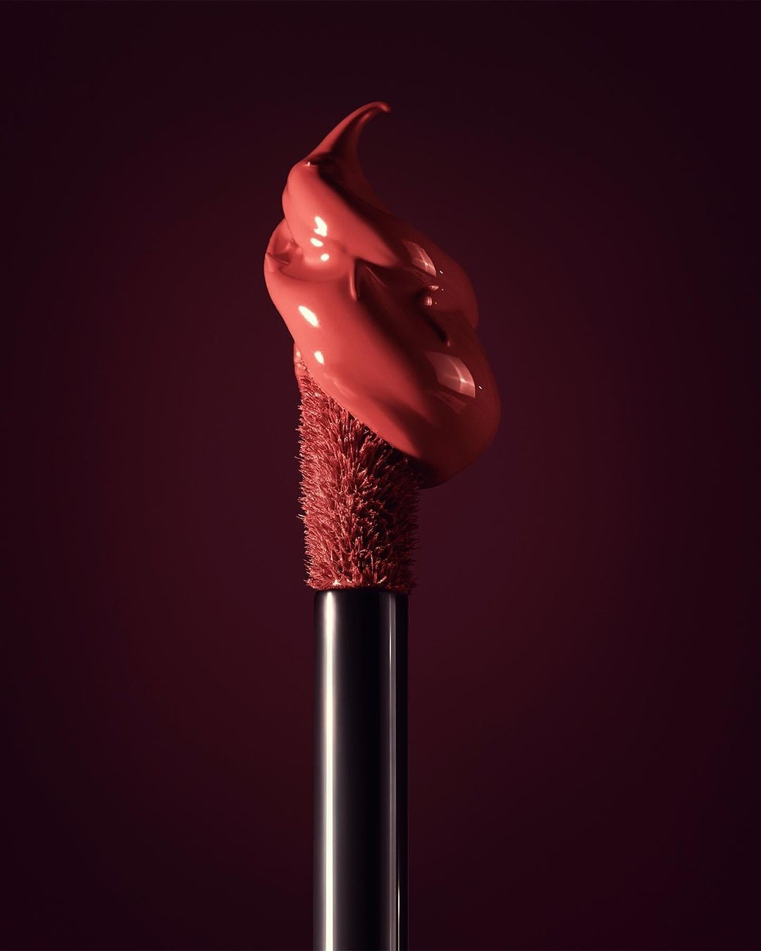 Close-up of a makeup applicator with a blob of coral-colored lip gloss on a deep red background, showcasing beauty and cosmetic product.