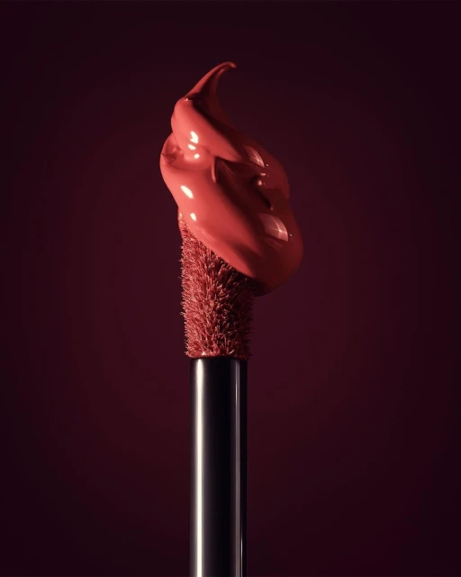 Close-up of a makeup applicator with a blob of coral-colored lip gloss on a deep red background, showcasing beauty and cosmetic product.