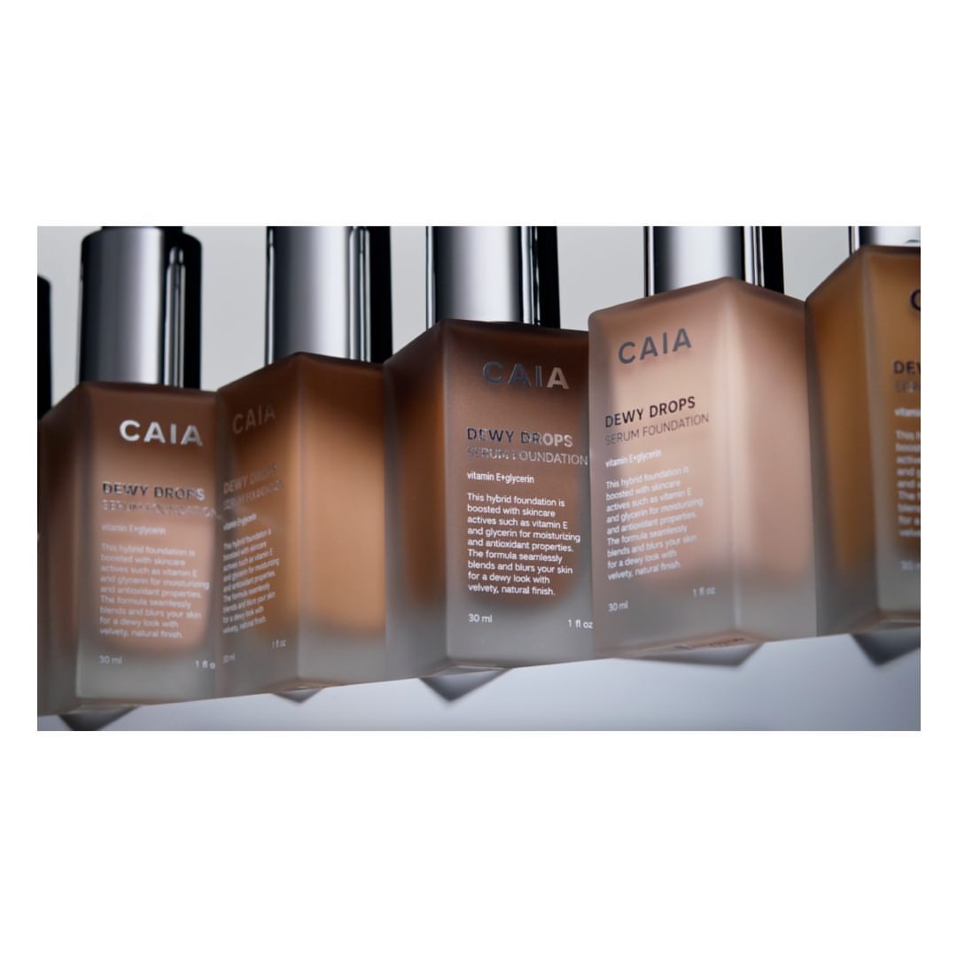 Several bottles of CAIA DeWy Drops serum foundation are displayed, showcasing the various shades and branding details in a studio setting.