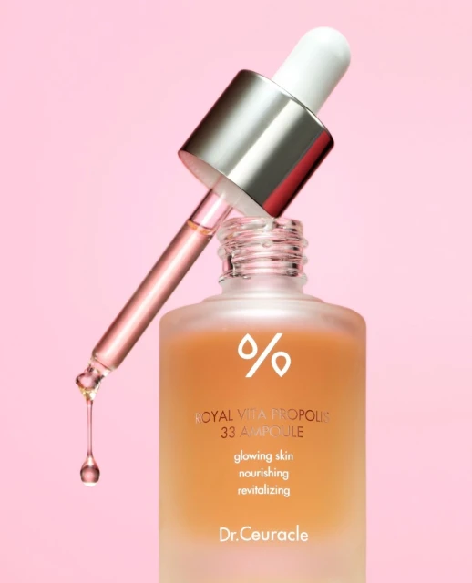 Close-up studio shot of Royal Vita Propolis 33 Ampoule by Dr. Ceuracle with a dropper poised above the bottle, on a pink background.