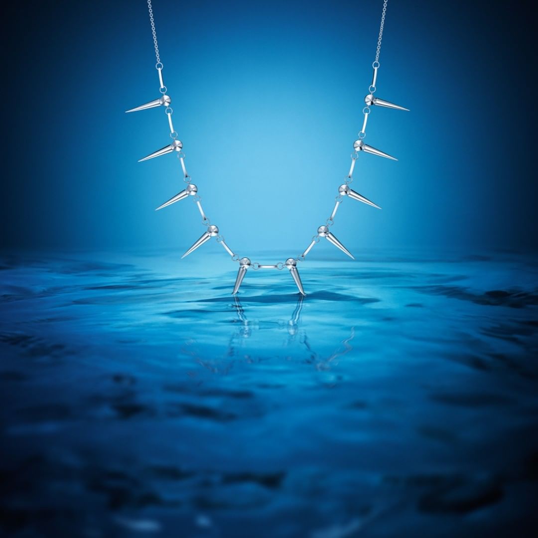 Close-up studio shot of a silver-colored necklace with spike accents, displayed against a water-like blue backdrop.