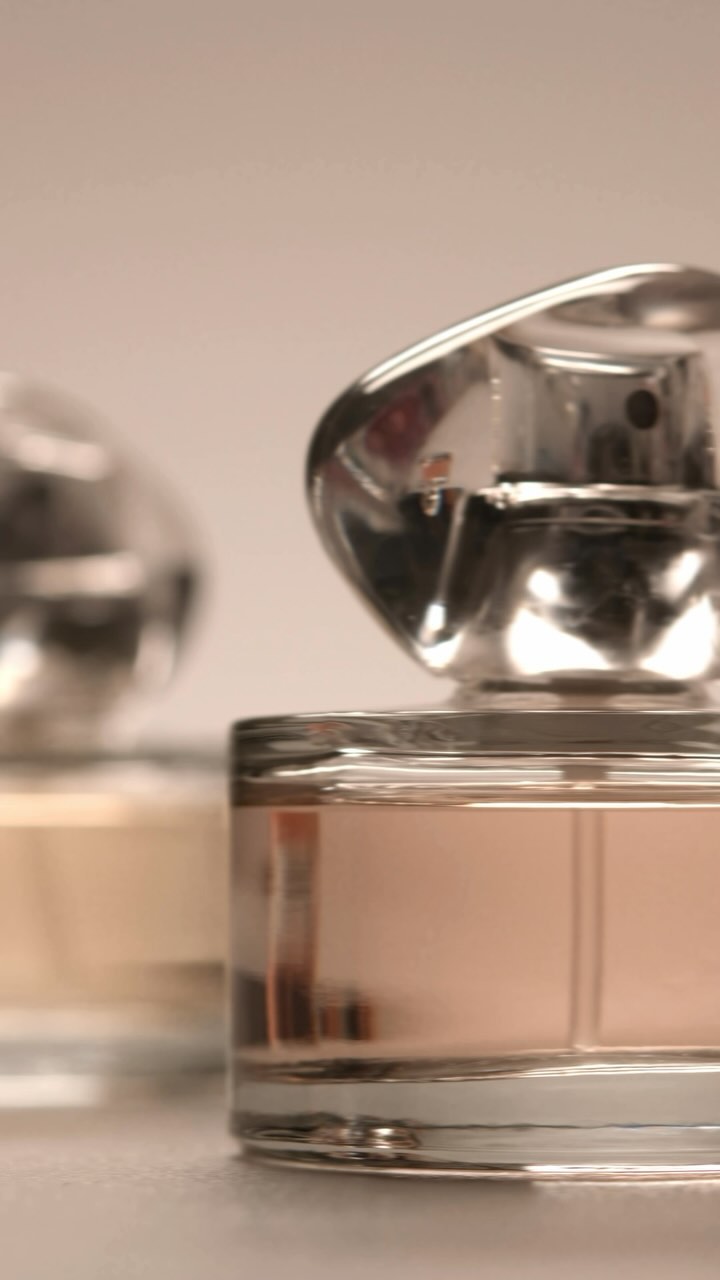 Two perfume bottles with clear glass and silver caps are displayed close-up against a soft, neutral background in vertical format.